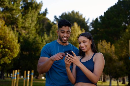 Téléchargez les photos : Heureux jeune couple multiracial portant des vêtements de sport souriant tout en utilisant le téléphone mobile dans le parc - en image libre de droit