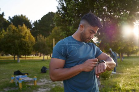 Téléchargez les photos : Jeune homme biracial portant un t-shirt regardant vers le bas le fitness tracker près des arbres dans le parc - en image libre de droit