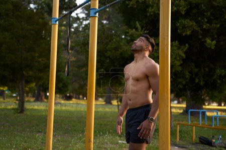 Photo for Shirtless young biracial man looking up at pull-up bar by trees in the park - Royalty Free Image
