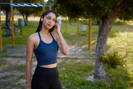 Photo for Young biracial woman wearing sportswear listening to music on wireless earphones in park - Royalty Free Image