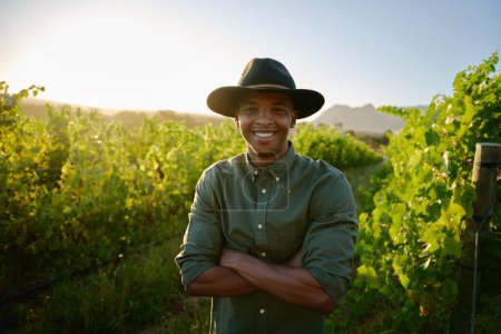 Téléchargez les photos : Heureux jeune homme noir portant un chapeau débordant souriant avec les bras croisés à côté des cultures à la ferme - en image libre de droit