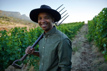 Foto de Feliz joven negro con ropa casual sosteniendo horquilla al lado de los cultivos en la granja - Imagen libre de derechos