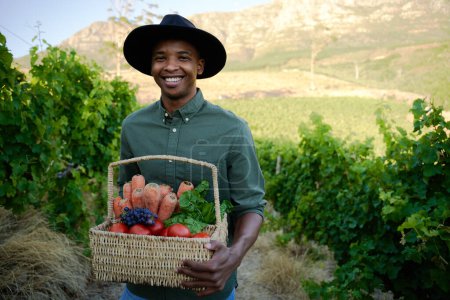 Foto de Joven hombre negro con ropa casual que sostiene la cesta con la cosecha de frutas y verduras en la granja - Imagen libre de derechos