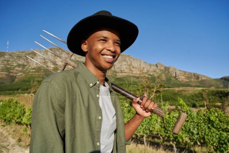 Photo for Happy young black man wearing shirt and brimmed hat holding pitchfork next to plants on farm - Royalty Free Image