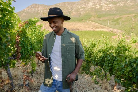 Foto de Joven hombre negro con camisa y sombrero de ala sonriendo mientras usa el teléfono al lado de las plantas en la granja - Imagen libre de derechos