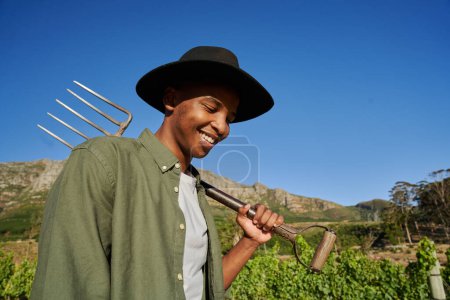Téléchargez les photos : Jeune homme noir portant chemise et chapeau débordé souriant tout en tenant fourche à côté de plantes à la ferme - en image libre de droit