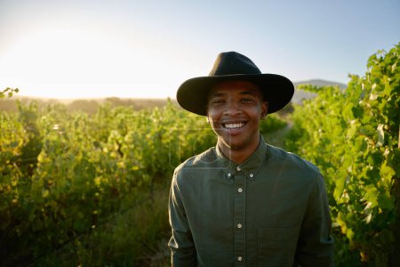 Foto de Retrato de un joven negro feliz con sombrero de ala sonriendo por los cultivos en la granja - Imagen libre de derechos