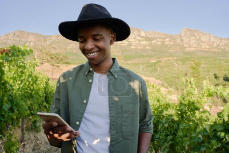 Téléchargez les photos : Jeune homme noir portant chemise et chapeau débordé souriant tout en utilisant le téléphone par les plantes à la ferme - en image libre de droit