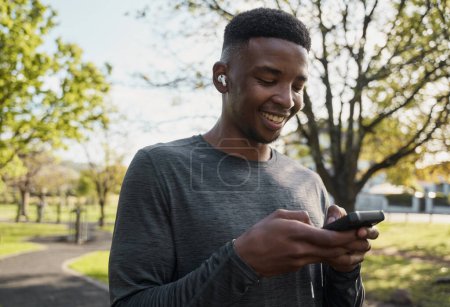 Téléchargez les photos : Heureux jeune homme noir portant des vêtements de sport avec écouteurs sans fil et téléphone portable dans le parc - en image libre de droit