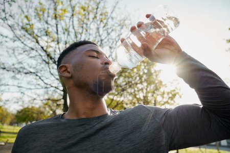 Foto de Joven hombre negro usando ropa deportiva con los ojos cerrados bebiendo agua de la botella en el parque - Imagen libre de derechos