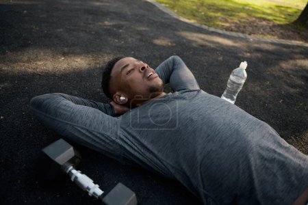 Foto de Joven hombre negro con ropa deportiva con auriculares inalámbricos acostado mientras toma un descanso en el parque - Imagen libre de derechos
