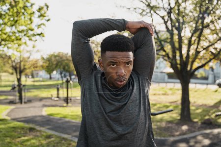 Foto de Joven hombre negro con ropa deportiva exhalando mientras hace estiramientos con los brazos levantados en el parque - Imagen libre de derechos
