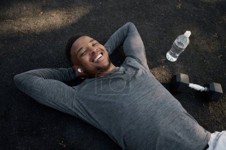 Photo for Happy young black man wearing sports clothing with wireless earphones lying down while resting in park - Royalty Free Image