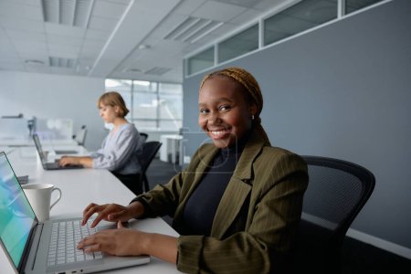 Téléchargez les photos : Deux jeunes femmes d'affaires multiraciales portant des vêtements d'affaires souriantes et tapant sur des ordinateurs portables au bureau - en image libre de droit