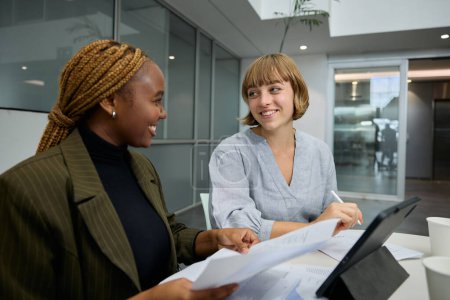 Téléchargez les photos : Deux jeunes femmes d'affaires multiraciales portant des vêtements d'affaires souriant par tablette numérique et des documents au bureau - en image libre de droit
