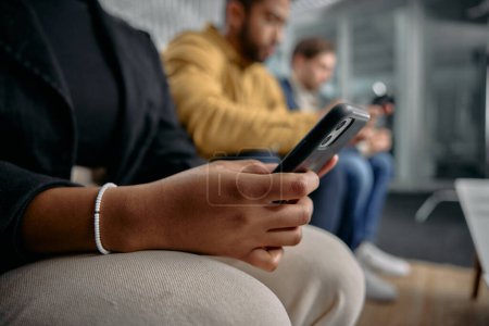 Photo for Three multiracial young adults wearing businesswear sitting and scrolling on mobile phones in office - Royalty Free Image