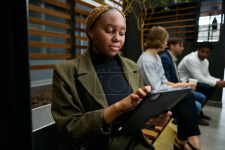 Téléchargez les photos : Quatre jeunes adultes concentrés portant des vêtements d'affaires parlant et utilisant une tablette numérique sur le banc dans le couloir - en image libre de droit