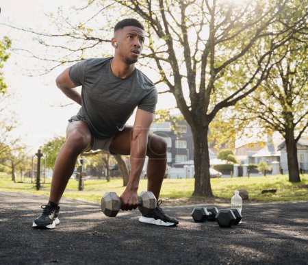 Photo for Fit young black man wearing sports clothing and wireless earphones doing squats with dumbbells in park - Royalty Free Image