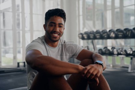 Téléchargez les photos : Heureux jeune homme multiracial portant des vêtements de sport assis et souriant tout en prenant une pause à la salle de gym - en image libre de droit