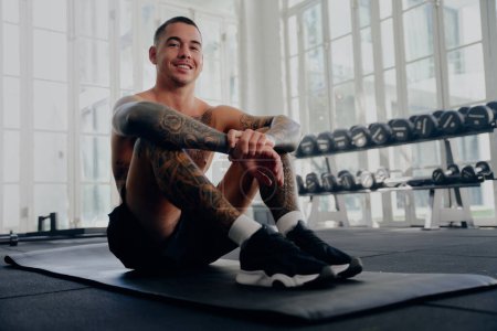 Foto de Shirtless joven tatuado hombre multirracial con pantalones cortos sonriendo y sentado mientras toma un descanso en el gimnasio - Imagen libre de derechos