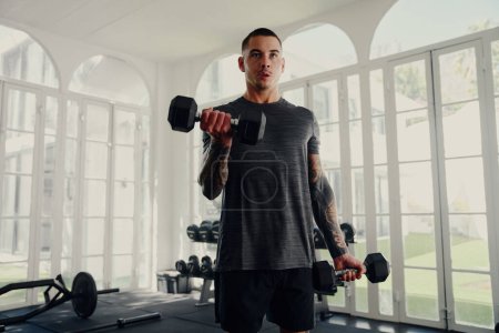 Foto de Joven hombre multirracial tatuado usando entrenamiento de peso de ropa deportiva en el gimnasio - Imagen libre de derechos
