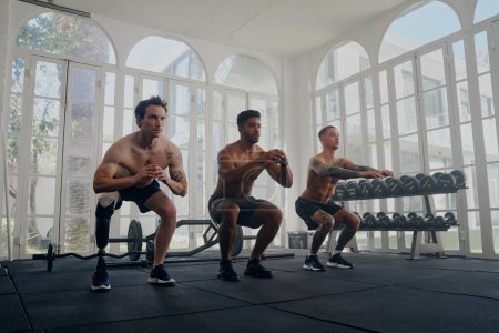 Téléchargez les photos : Trois jeunes hommes multiraciaux torse nu portant des shorts faisant squats dans une rangée à la salle de gym - en image libre de droit