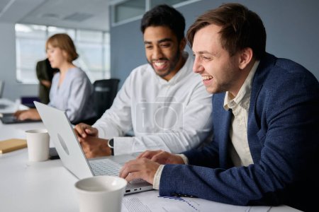 Téléchargez les photos : Quatre jeunes gens d'affaires multiraciaux heureux portant des vêtements d'affaires travaillant sur des ordinateurs portables au bureau - en image libre de droit