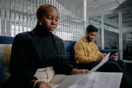 Téléchargez les photos : Deux jeunes adultes multiraciaux portant des documents de lecture de vêtements d'affaires sur le canapé dans le bureau d'entreprise - en image libre de droit