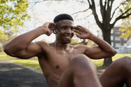 Foto de Shirtless joven negro con auriculares inalámbricos haciendo abdominales mientras sonríe en el parque - Imagen libre de derechos