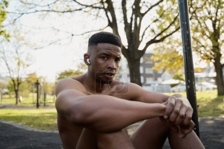 Photo for Shirtless young black man wearing wireless earphones sitting and looking at camera in park - Royalty Free Image