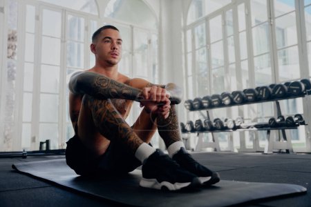 Foto de Shirtless joven tatuado multirracial hombre con pantalones cortos sentado mientras toma un descanso en el gimnasio - Imagen libre de derechos