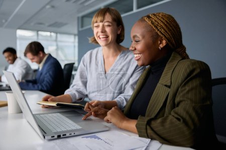 Téléchargez les photos : Quatre jeunes hommes d'affaires multiraciaux portant des vêtements d'affaires souriant tout en travaillant sur des ordinateurs portables au bureau - en image libre de droit