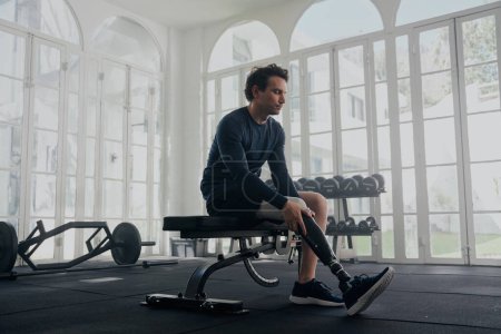 Foto de Joven hombre caucásico con ropa deportiva sentado en press de banca mientras se adjunta la pierna protésica en el gimnasio - Imagen libre de derechos