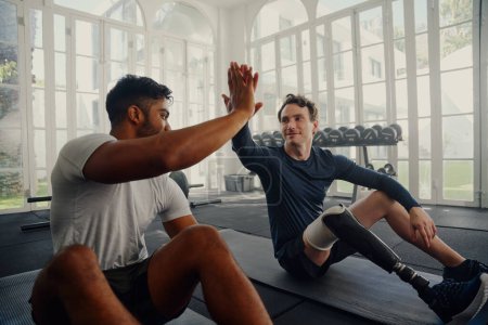 Photo for Two young multiracial men wearing sports clothing sitting on exercise mats and high-fiving at the gym - Royalty Free Image