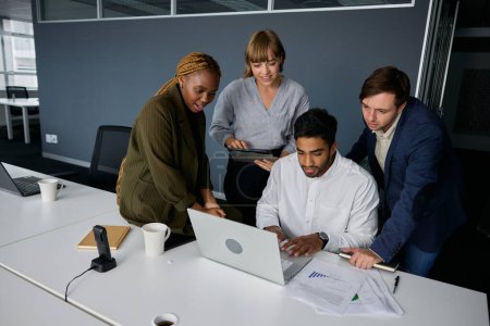 Téléchargez les photos : Quatre jeunes adultes portant des vêtements d'affaires parlant et utilisant une tablette numérique et un ordinateur portable sur le bureau - en image libre de droit