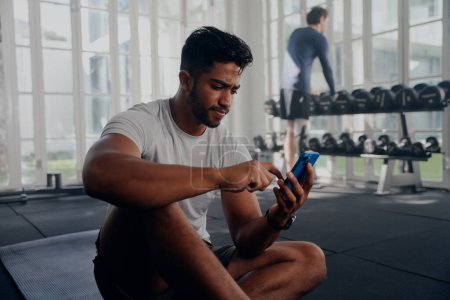 Photo for Two multiracial young men wearing sportswear using mobile phone next to weights at the gym - Royalty Free Image