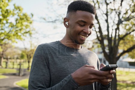 Foto de Feliz joven negro con ropa deportiva con auriculares inalámbricos y teléfono móvil en el parque - Imagen libre de derechos