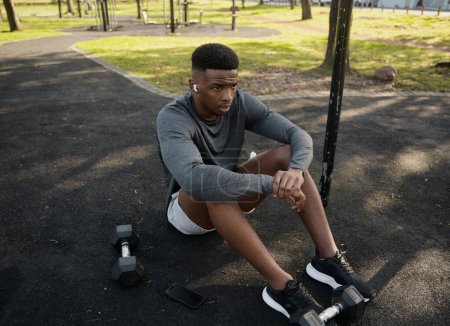 Foto de Joven hombre negro con ropa deportiva con auriculares inalámbricos sentado tomando un descanso en el parque - Imagen libre de derechos