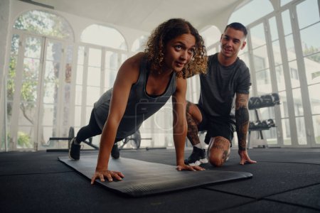 Foto de Entrenador multirracial con una mujer joven vestida con ropa deportiva haciendo ejercicios push-up en el gimnasio - Imagen libre de derechos
