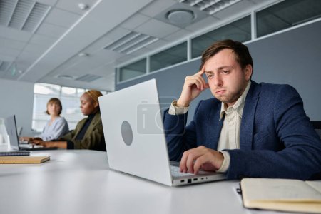 Foto de Tres jóvenes empresarios multirraciales estresados que usan ropa de negocios trabajando en el escritorio en computadoras portátiles en la oficina - Imagen libre de derechos