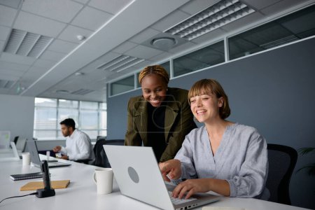 Photo for Three young multiracial business people wearing businesswear smiling and typing on laptop at desk in office - Royalty Free Image