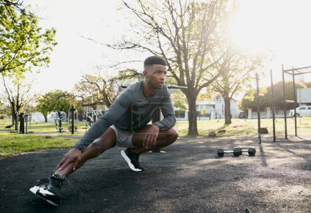 Photo for Young black man wearing sports clothing doing stretches in squatting position next to dumbbells at park - Royalty Free Image