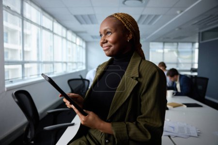 Foto de Joven empresaria con ropa de negocios sosteniendo tableta digital mientras está sentada en el escritorio en la oficina corporativa - Imagen libre de derechos
