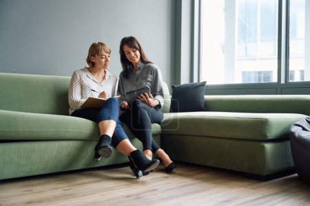 Téléchargez les photos : Deux femmes d'affaires assises sur un canapé souriant en utilisant une tablette numérique lors d'une réunion dans un bureau d'entreprise - en image libre de droit