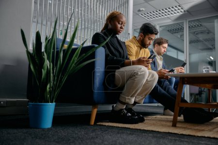 Photo for Three multiracial young adults wearing businesswear sitting on sofa and using mobile phones in office - Royalty Free Image