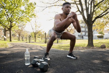 Shirtless young black man wearing wireless earphones doing squats next to dumbbells in park