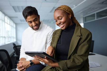 Foto de Jóvenes empresarios felices vistiendo ropa de negocios sentados en el escritorio mientras usan tabletas digitales en la oficina corporativa - Imagen libre de derechos