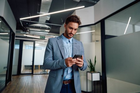 Téléchargez les photos : Jeune homme caucasien portant des vêtements d'affaires en utilisant le téléphone mobile dans le couloir du bureau d'entreprise - en image libre de droit