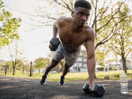 Téléchargez les photos : Jeune homme noir torse nu portant des écouteurs sans fil faisant de l'exercice avec des haltères dans le parc - en image libre de droit