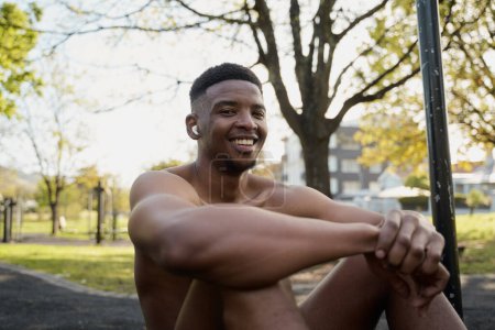 Téléchargez les photos : Jeune homme noir torse nu portant des écouteurs sans fil assis et souriant dans le parc - en image libre de droit